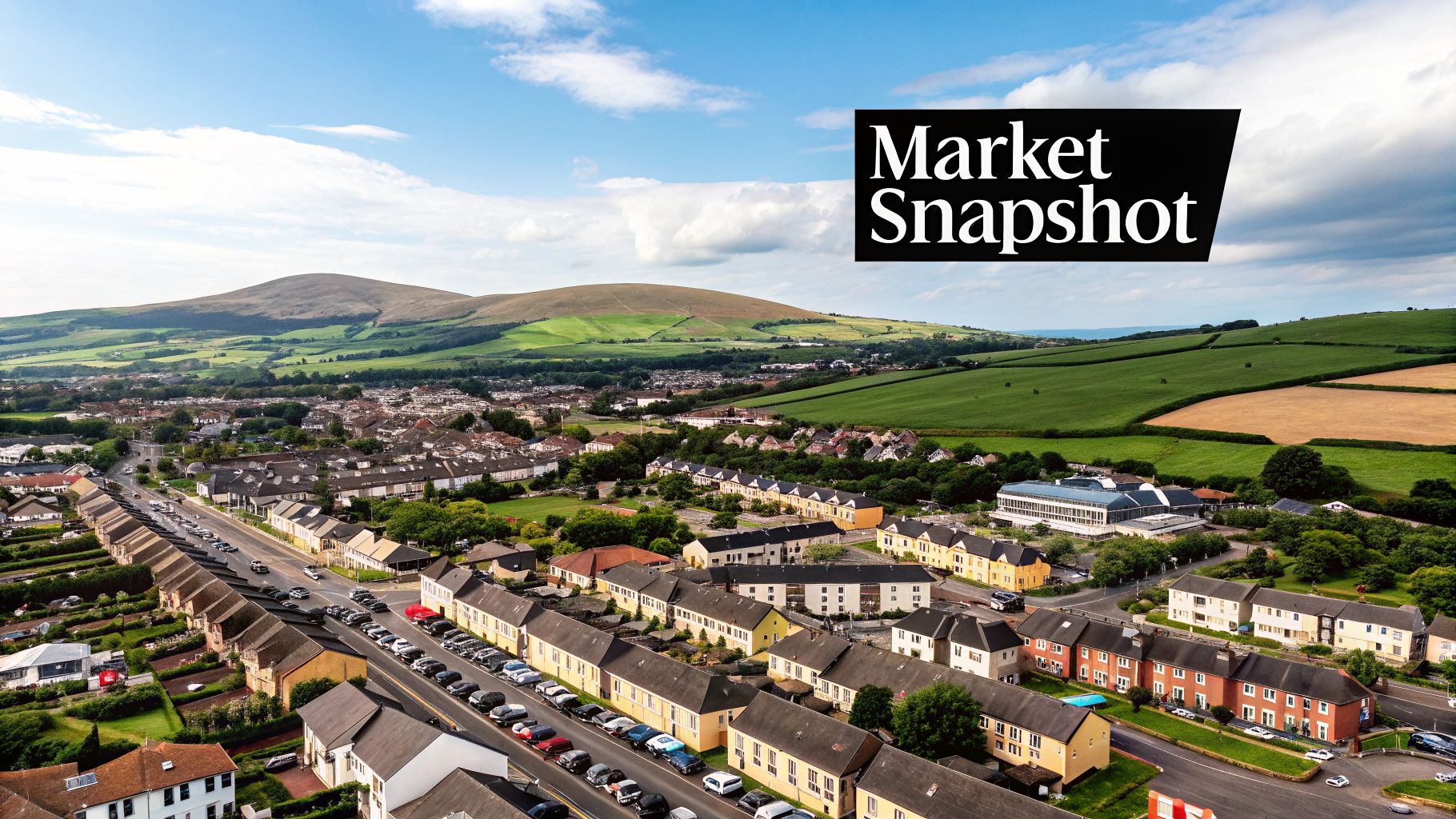 Aerial view of a residential town with rows of houses, cars, green hills, and fields under a sunny sky.