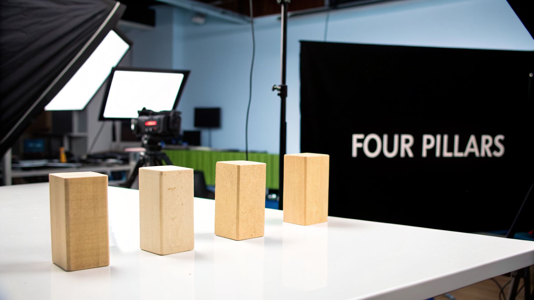 Four wooden blocks stand on a white table in a studio, with lights, a camera, and a 'FOUR PILLARS' sign in the background.
